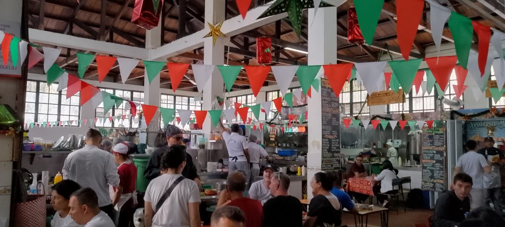 the inside of la perseverancia market. people sit at tables and there are red, white, and green flags strewn from different parts of the building